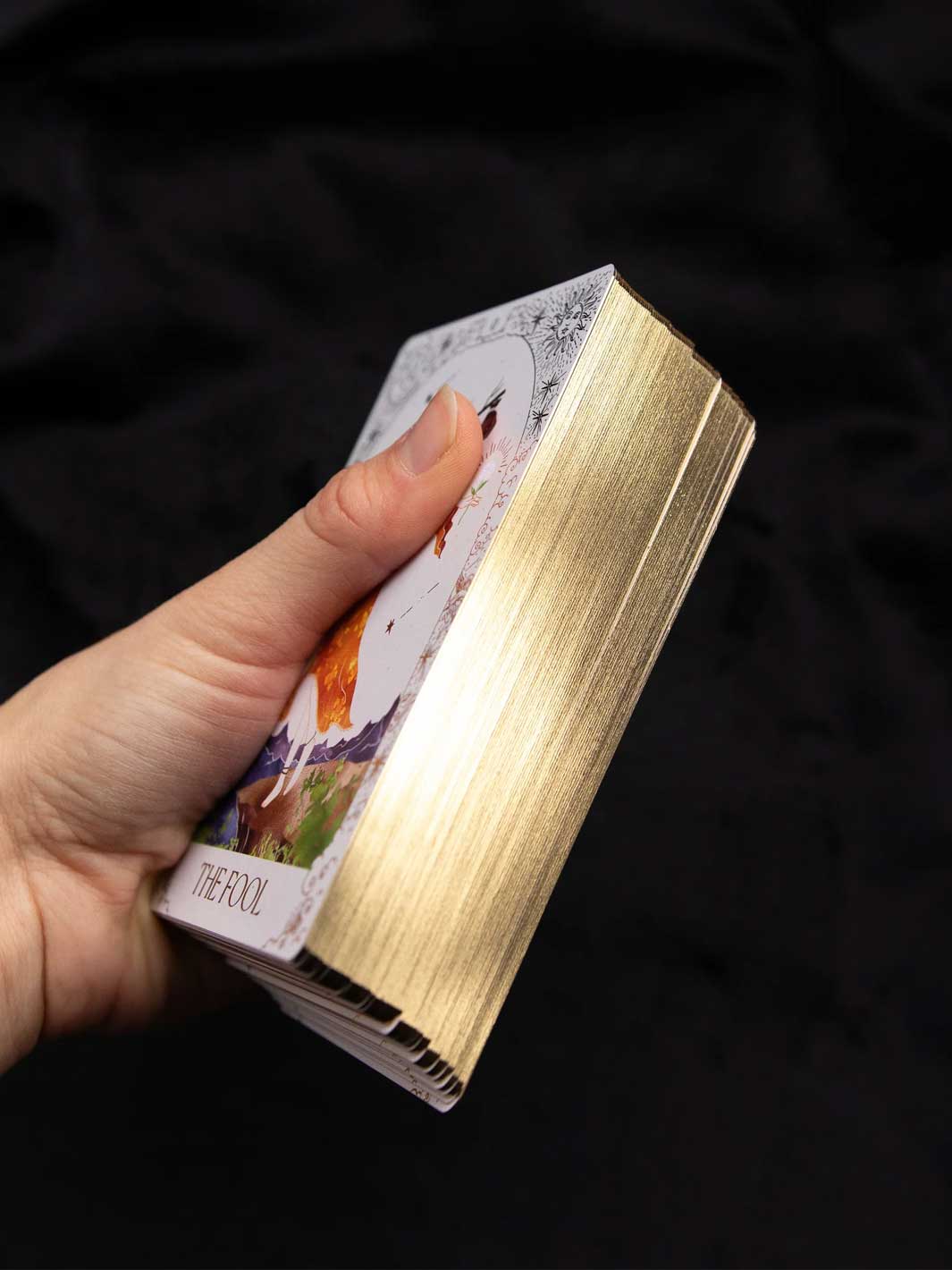 A hand holding a deck of tarot cards with a metallic gold-leaf textured spine and a white card face visible, set against a dark black fabric background.