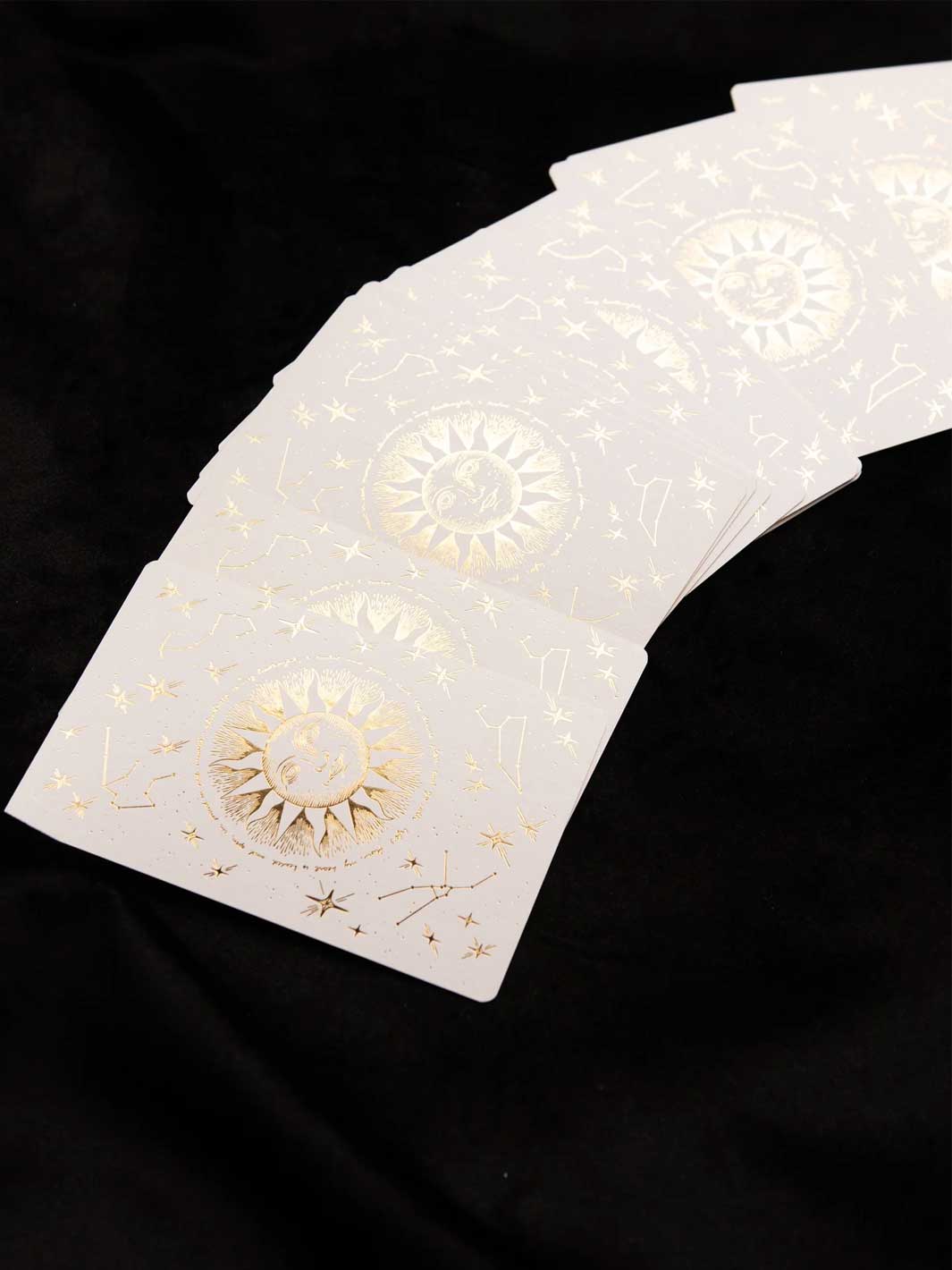 A fanned-out deck of white oracle cards decorated with gold foil celestial designs, including suns, moons, and constellations, set against a black fabric background.