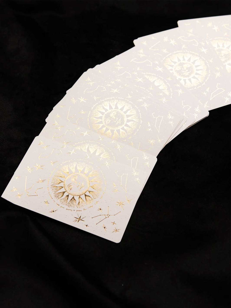A fanned-out deck of white oracle cards decorated with gold foil celestial designs, including suns, moons, and constellations, set against a black fabric background.