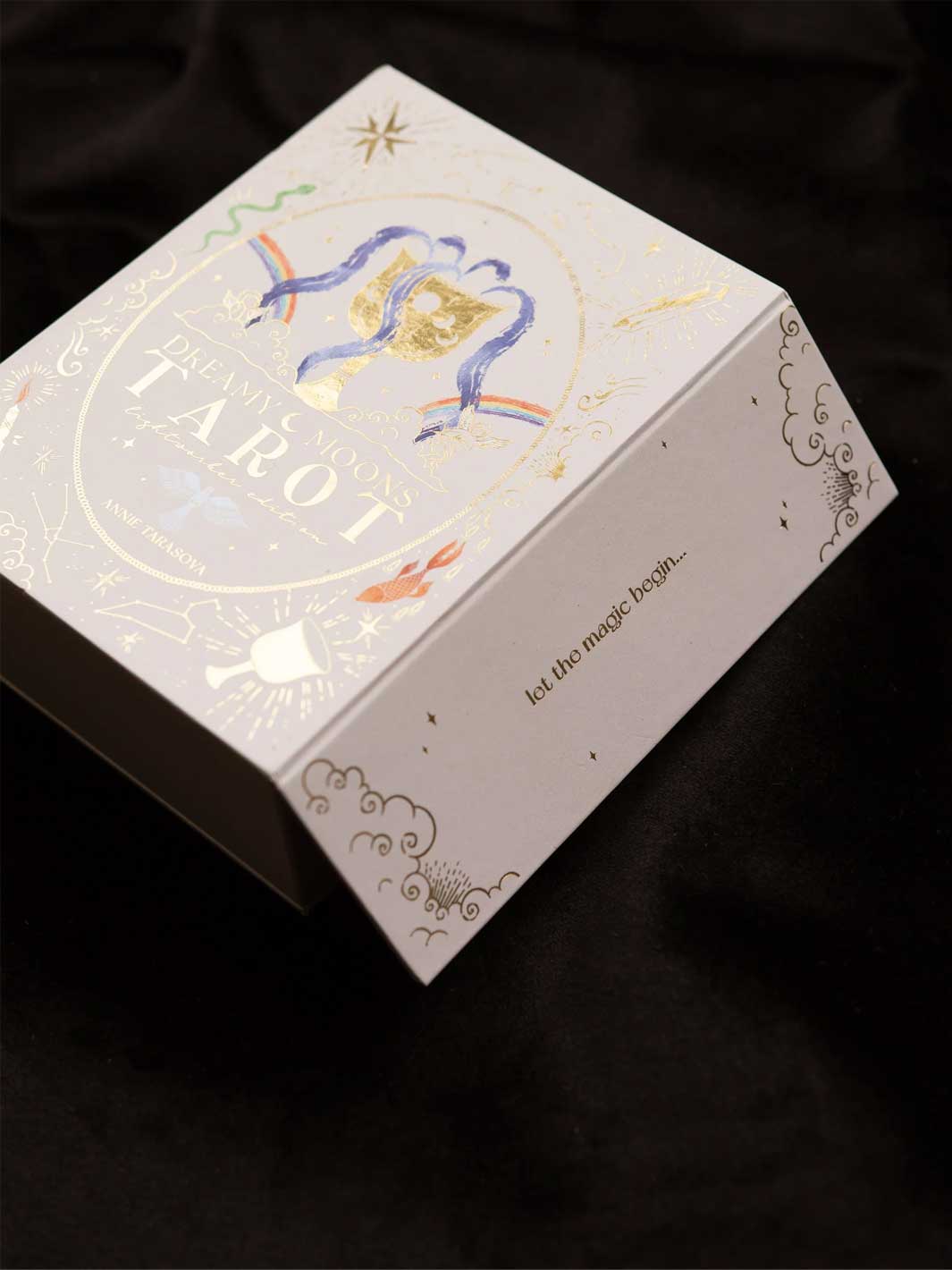 A white tarot card box with elegant gold foil details, featuring the title 'Dreamy Moons Tarot' and a blue and gold illustration of two birds, set against a dark black fabric background.