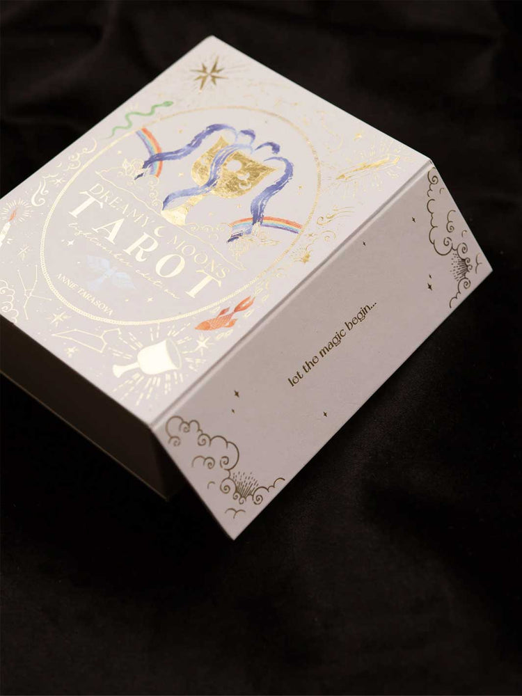 A white tarot card box with elegant gold foil details, featuring the title 'Dreamy Moons Tarot' and a blue and gold illustration of two birds, set against a dark black fabric background.