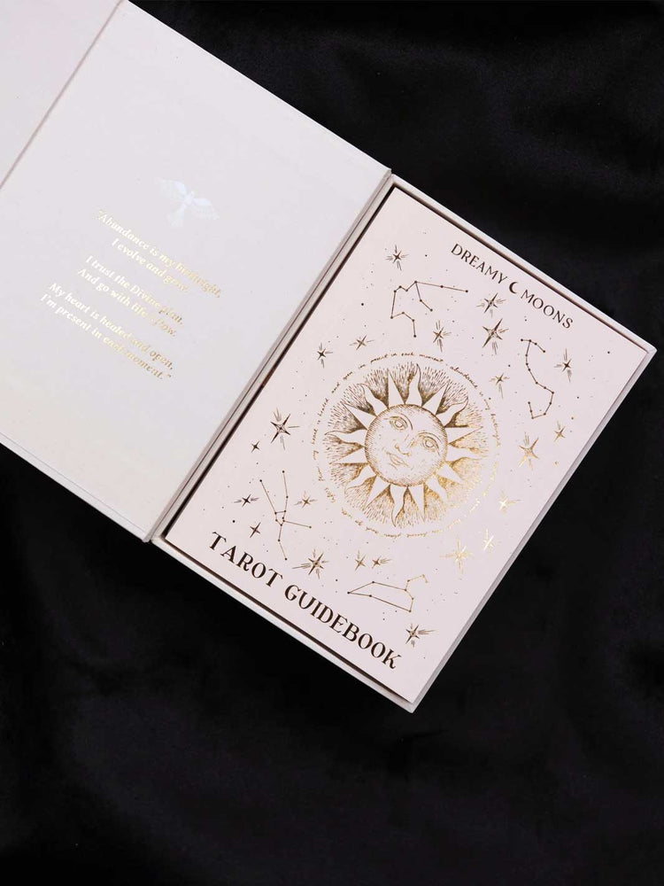 A tarot guidebook titled 'Dreamy Moon's Tarot Guidebook' displayed open on a black fabric background. The cover features a cream-colored background with gold foil illustrations of a sun, stars, and constellations.