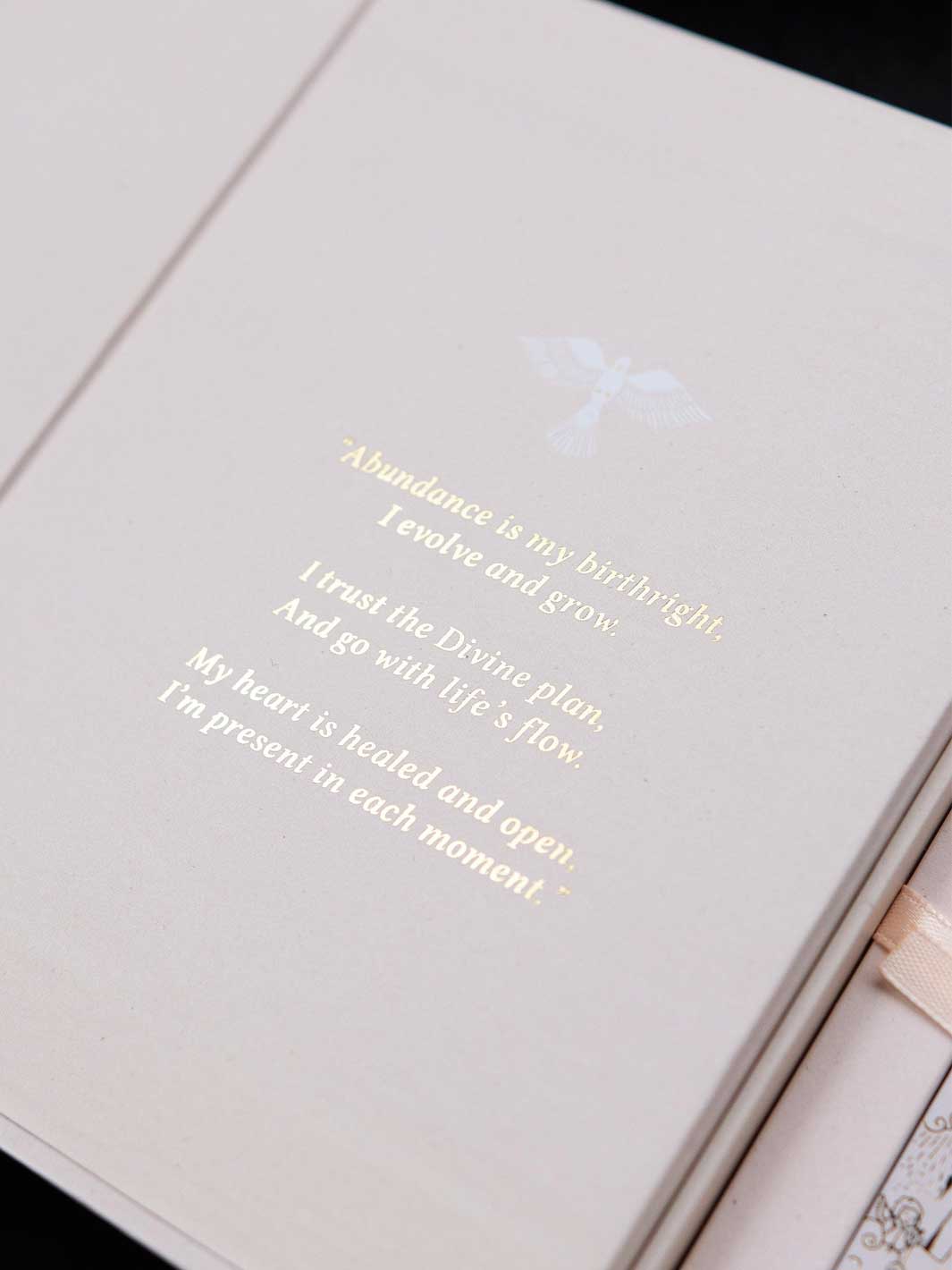 A close-up of a cream-colored journal or notebook page with elegant gold foil text and a small angel illustration. The text reads, &
