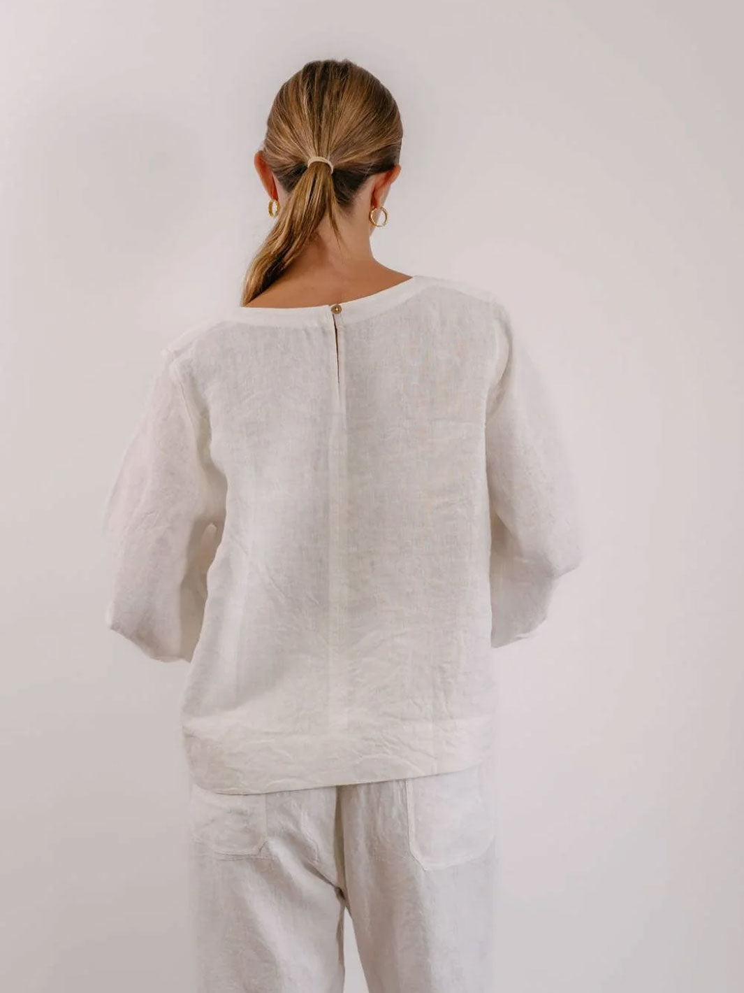 A woman viewed from behind wearing a loose white linen top and matching white pants against a plain light background.