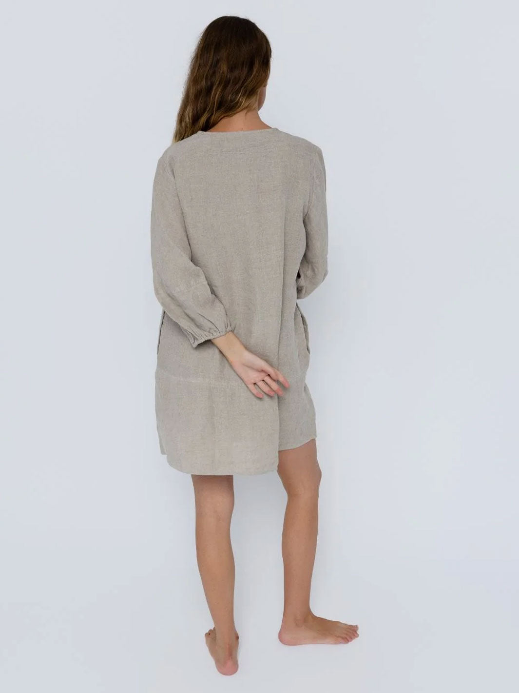 A woman viewed from behind wearing a light beige linen long-sleeve tunic dress with side pockets, standing against a plain light gray background.