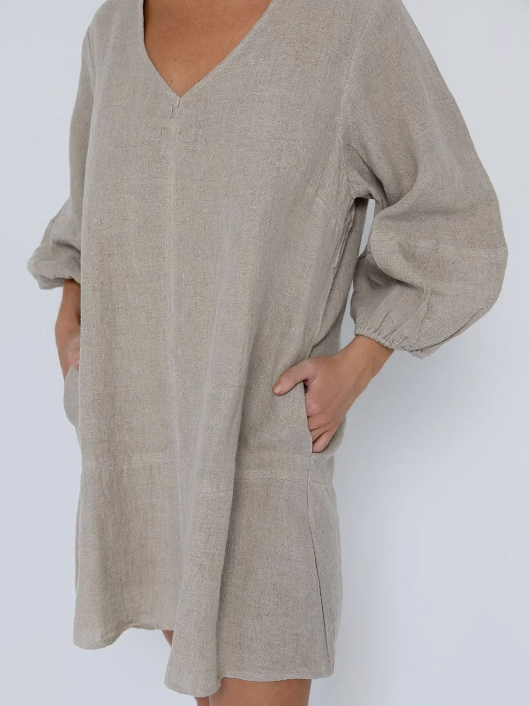 A person wearing a loose, oversized linen dress or tunic in a light taupe-beige color, featuring a V-neckline, voluminous elbow-length sleeves, and side pockets, shown against a plain light gray background.