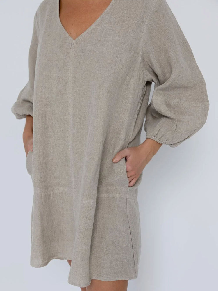 A person wearing a loose, oversized linen dress or tunic in a light taupe-beige color, featuring a V-neckline, voluminous elbow-length sleeves, and side pockets, shown against a plain light gray background.