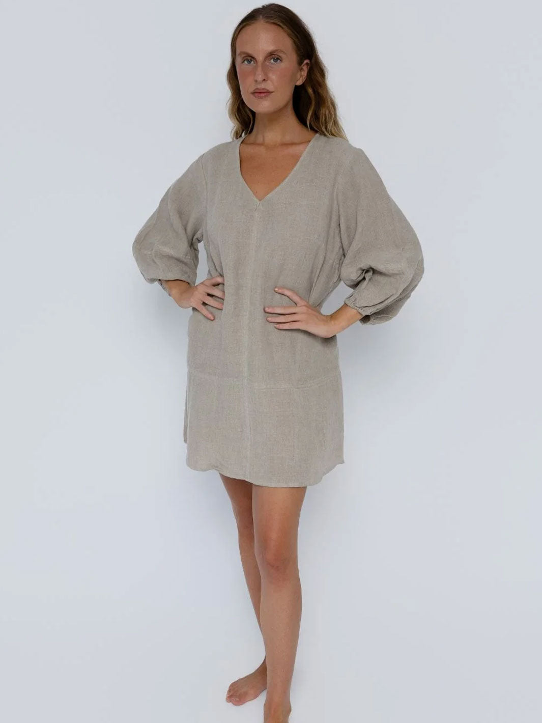 A woman modeling a light beige linen tunic dress with a V-neckline and voluminous balloon sleeves, standing against a plain light gray studio background.