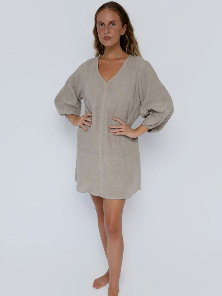 A woman modeling a light beige linen tunic dress with a V-neckline and voluminous balloon sleeves, standing against a plain light gray studio background.