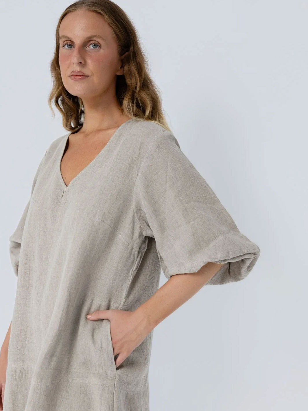 A woman wearing a loose, light beige linen tunic dress with a V-neck and elbow-length sleeves, posed against a light gray studio background.