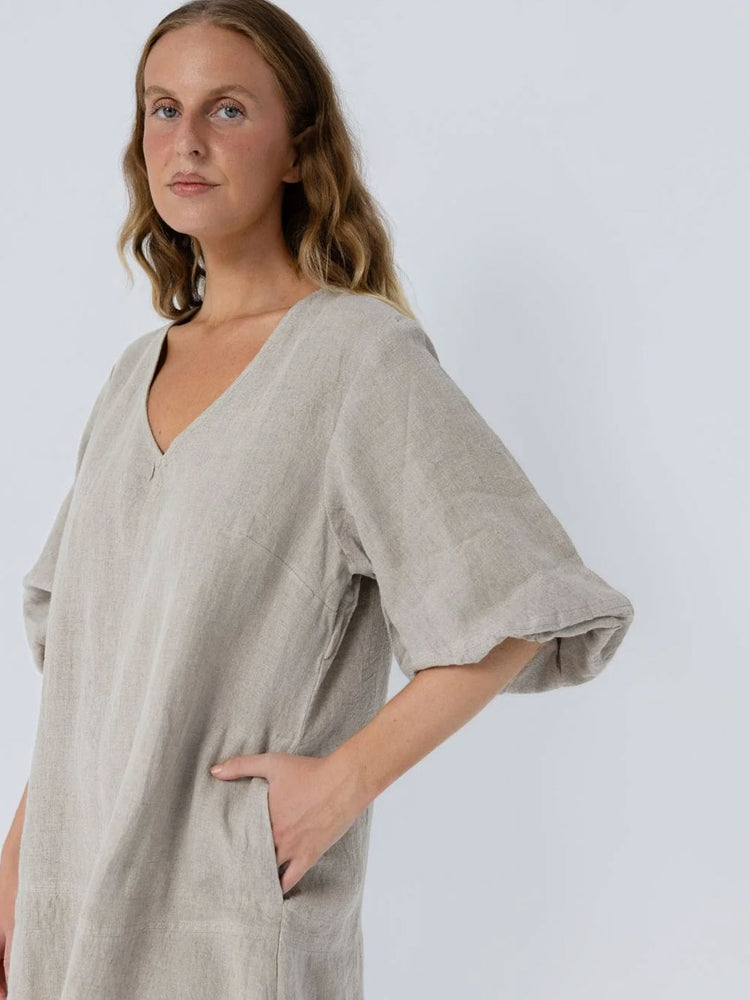 A woman wearing a loose, light beige linen tunic dress with a V-neck and elbow-length sleeves, posed against a light gray studio background.