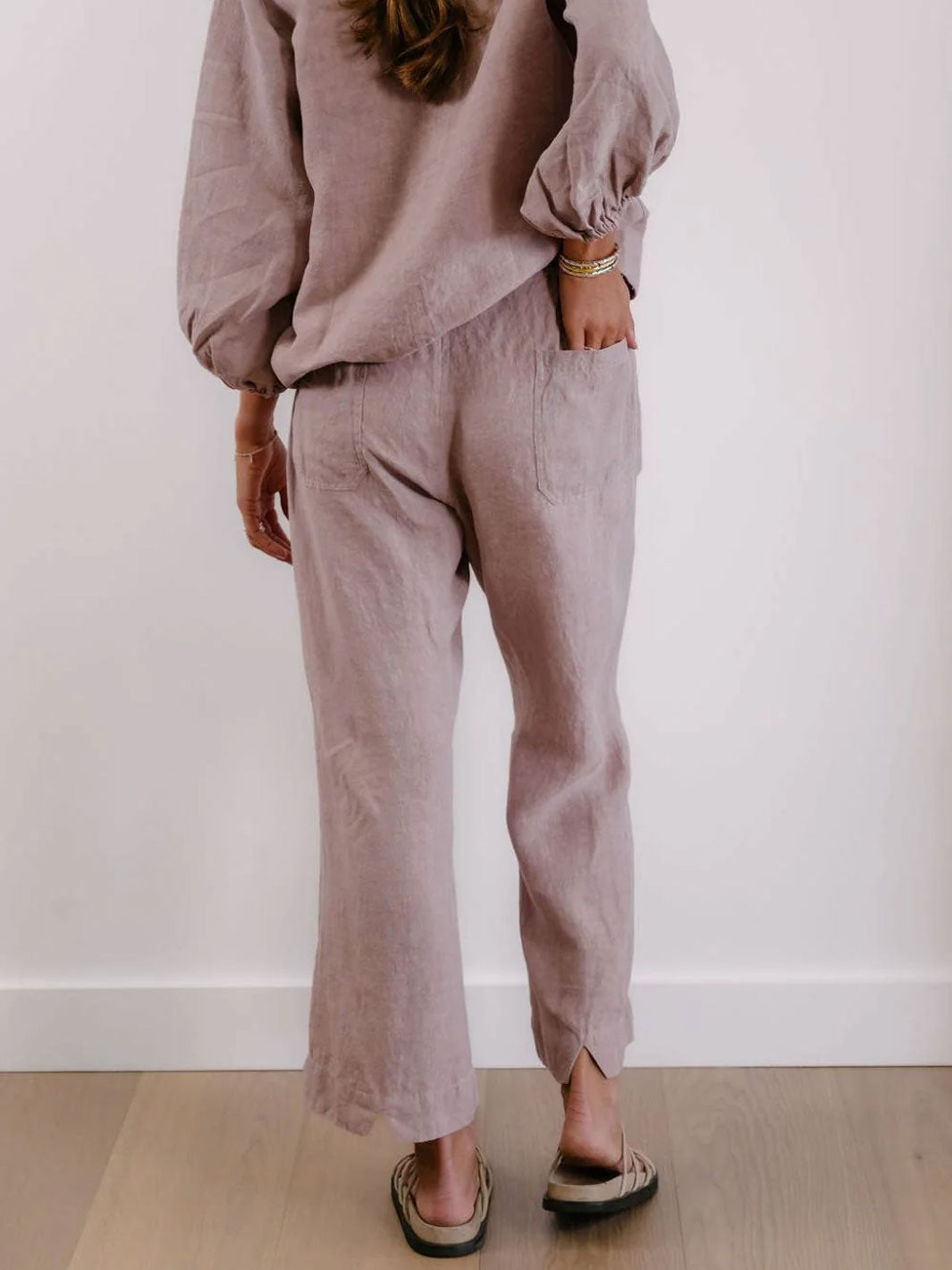 A person shown from the back wearing a matching dusty mauve linen set consisting of a long-sleeve top and wide-leg cropped pants, paired with dark sandals against a white wall and light wood floor.