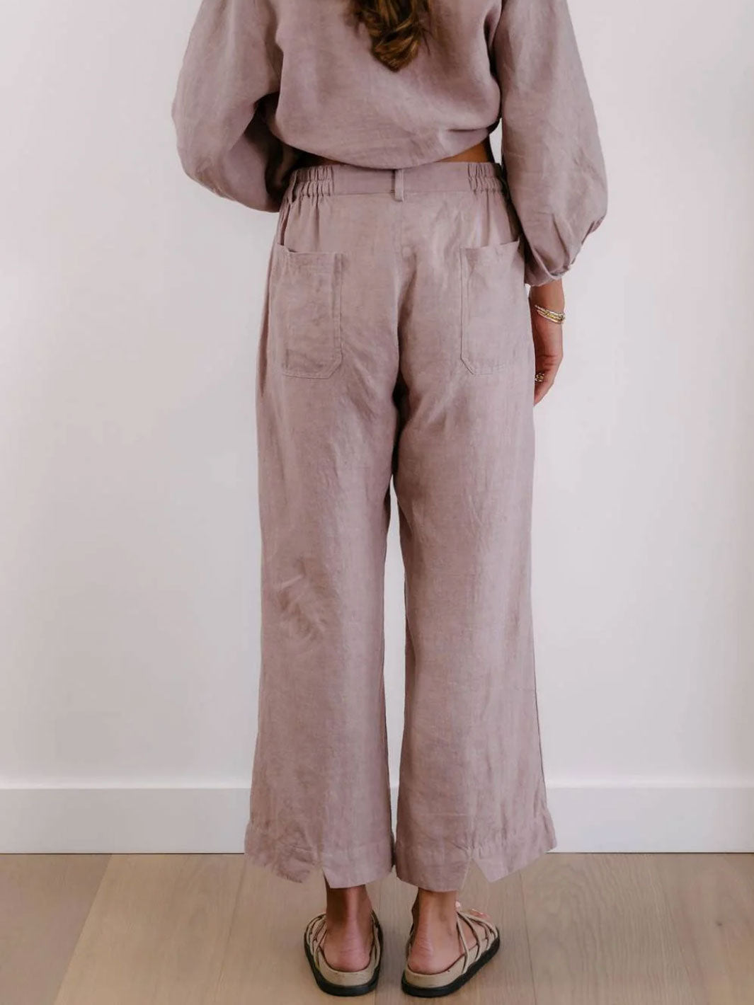 A person shown from the back wearing dusty mauve wide-leg linen pants with a matching long-sleeve top, standing on a light wood floor against a white wall.