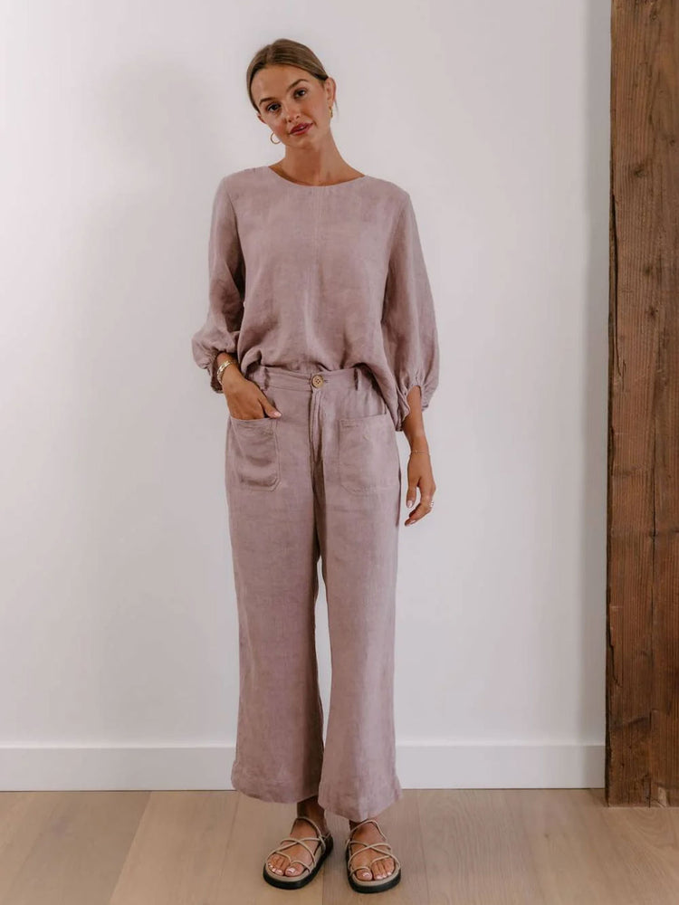 A woman modeling a matching dusty rose linen two-piece set consisting of a long-sleeve top and wide-leg trousers, paired with brown sandals, standing against a white wall with a wooden column on the right.
