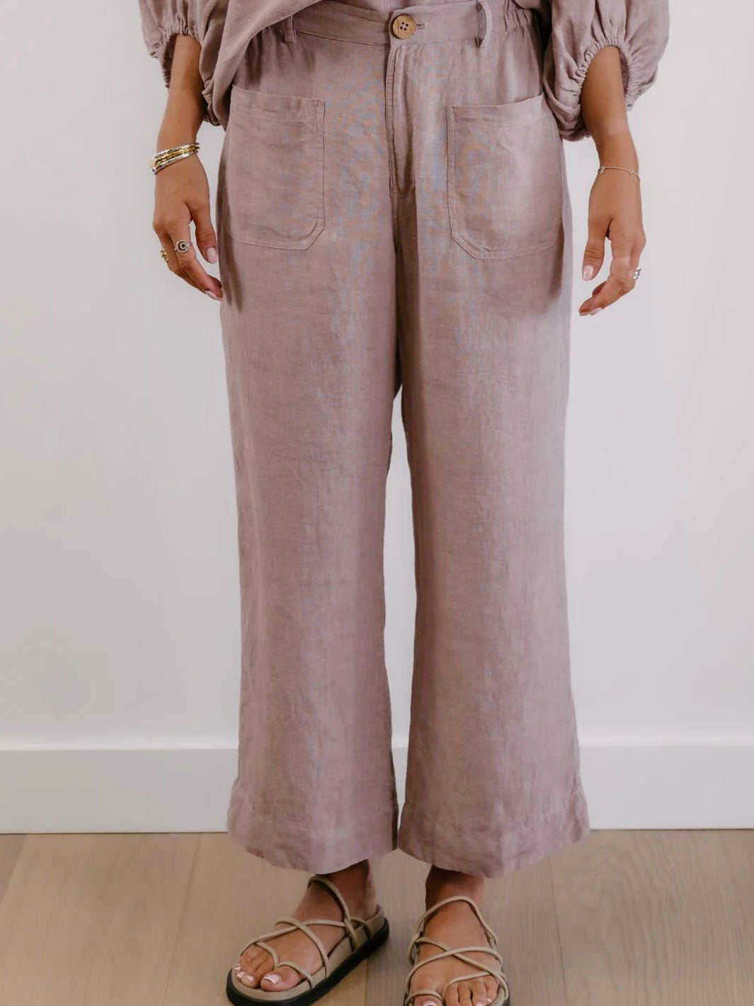 A person wearing dusty mauve wide-leg linen pants with front patch pockets, paired with a matching top and strappy flat sandals, standing against a white wall and light wood floor.