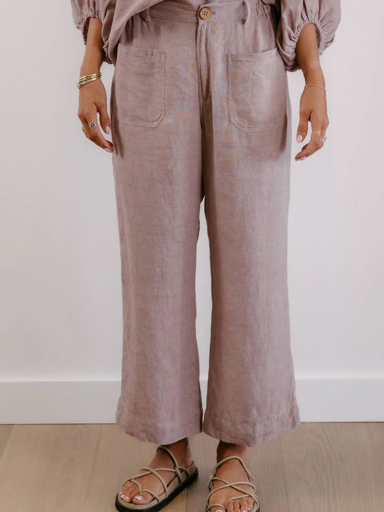 A person wearing dusty mauve wide-leg linen pants with front patch pockets, paired with a matching top and strappy flat sandals, standing against a white wall and light wood floor.