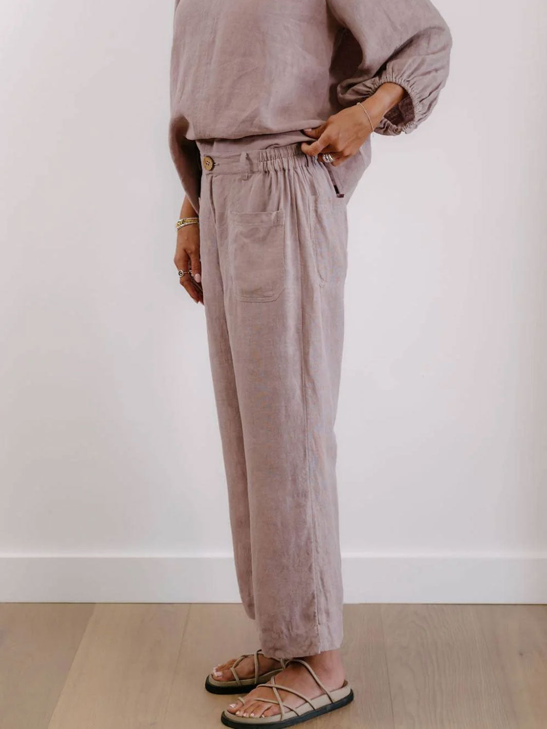A person wearing loose, high-waisted mauve linen trousers with a matching top and sandals, standing against a plain white wall on a light wood floor.