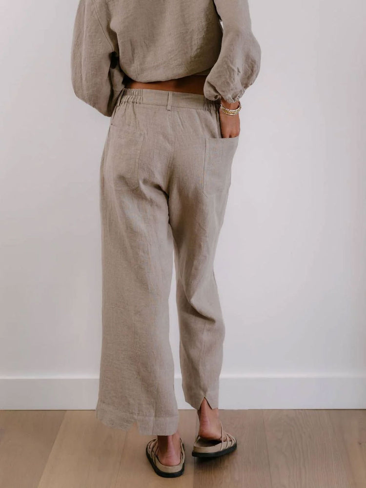 A person shown from the back wearing loose, wide-leg taupe linen pants with side pockets, paired with a matching beige top and sandals, standing against a white wall and light wood floor.