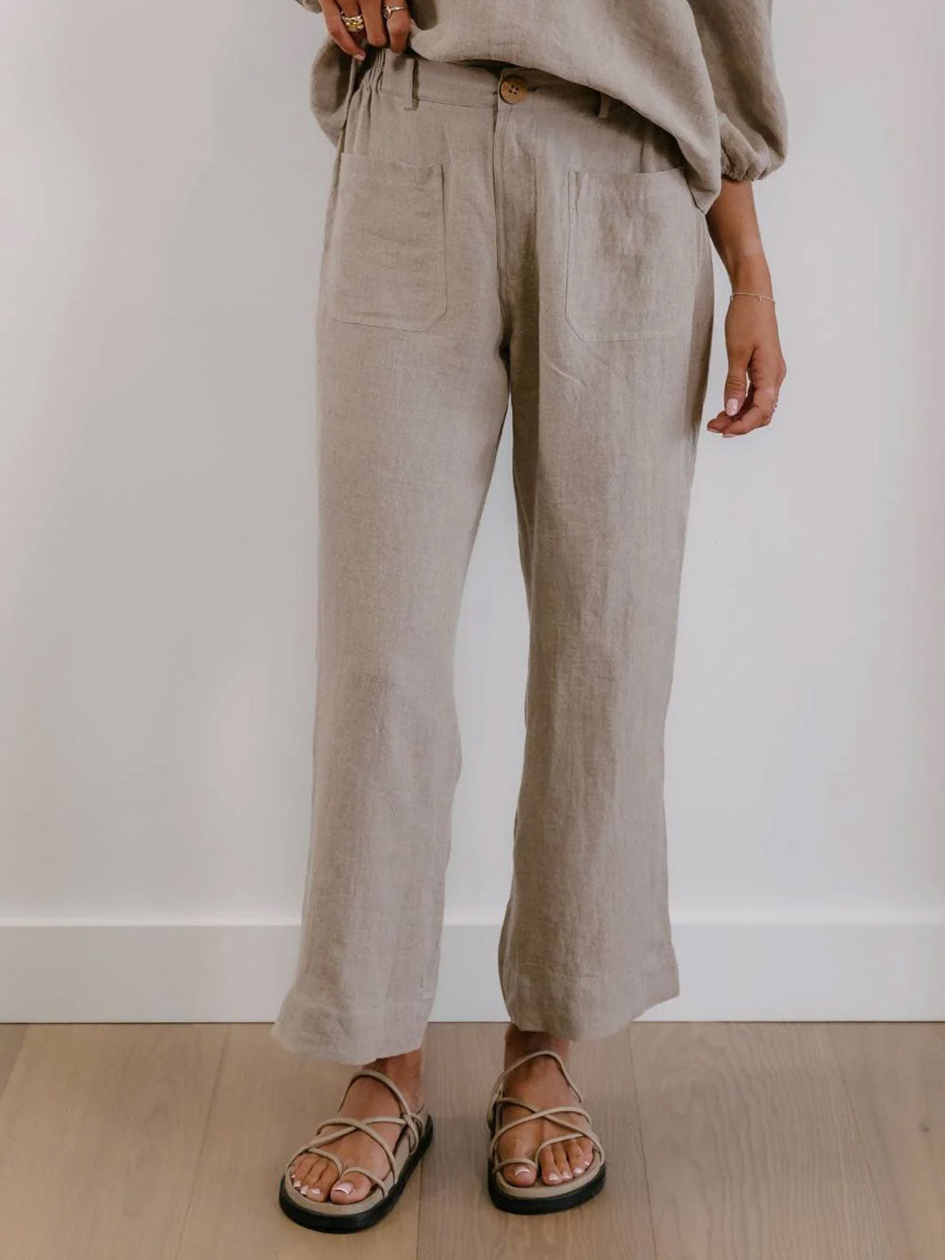 A person wearing loose, wide-leg linen pants in a light taupe-beige color, paired with a matching top and flat sandals, standing against a white wall and light wood floor.