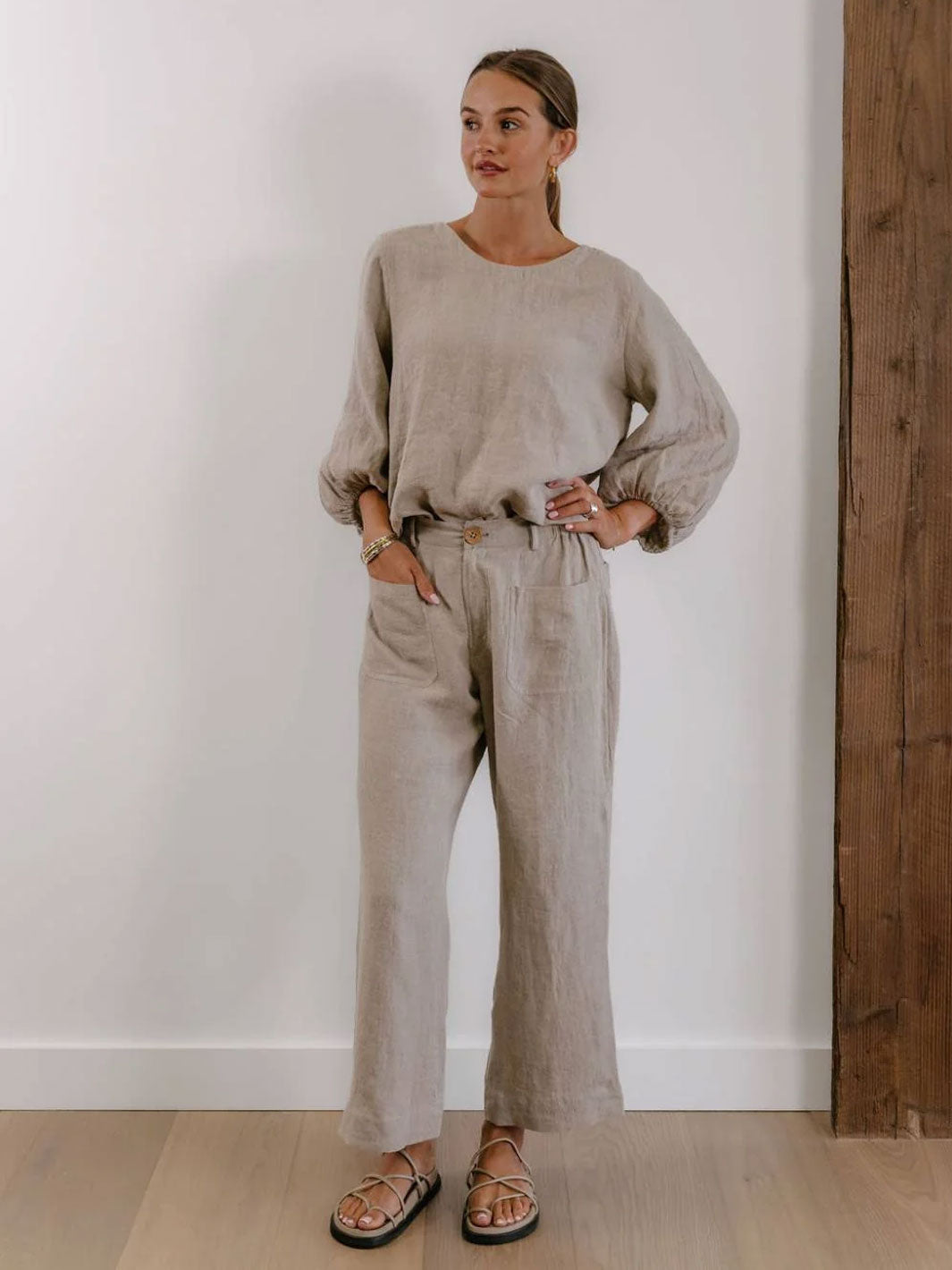 A woman modeling a light beige linen two-piece set consisting of a long-sleeve top and matching wide-leg trousers, paired with brown sandals, standing in a room with a white wall and wooden floor.