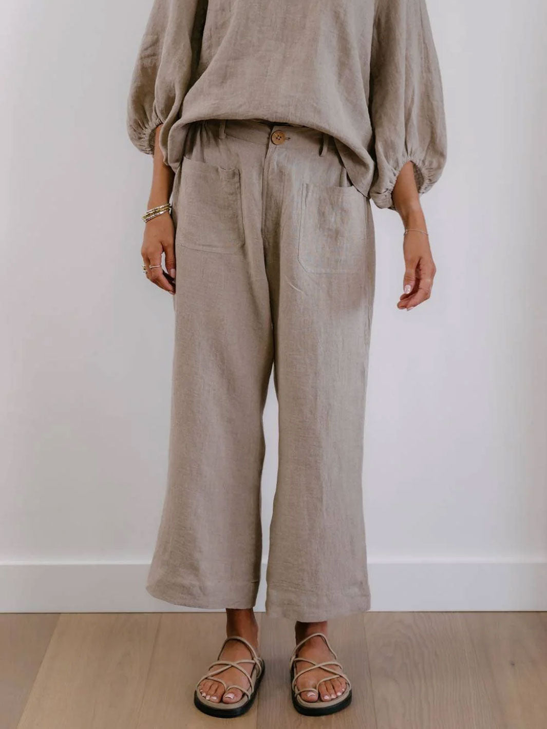 A person wearing loose, wide-leg linen pants in a light taupe-beige color, paired with a matching beige top and black strappy flat sandals, standing against a plain white wall.