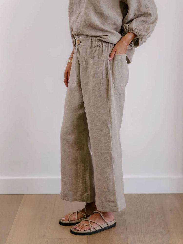 A person wearing loose, wide-leg linen pants in a light taupe-beige color, paired with a matching top and flat sandals, standing against a white wall on a light wood floor.