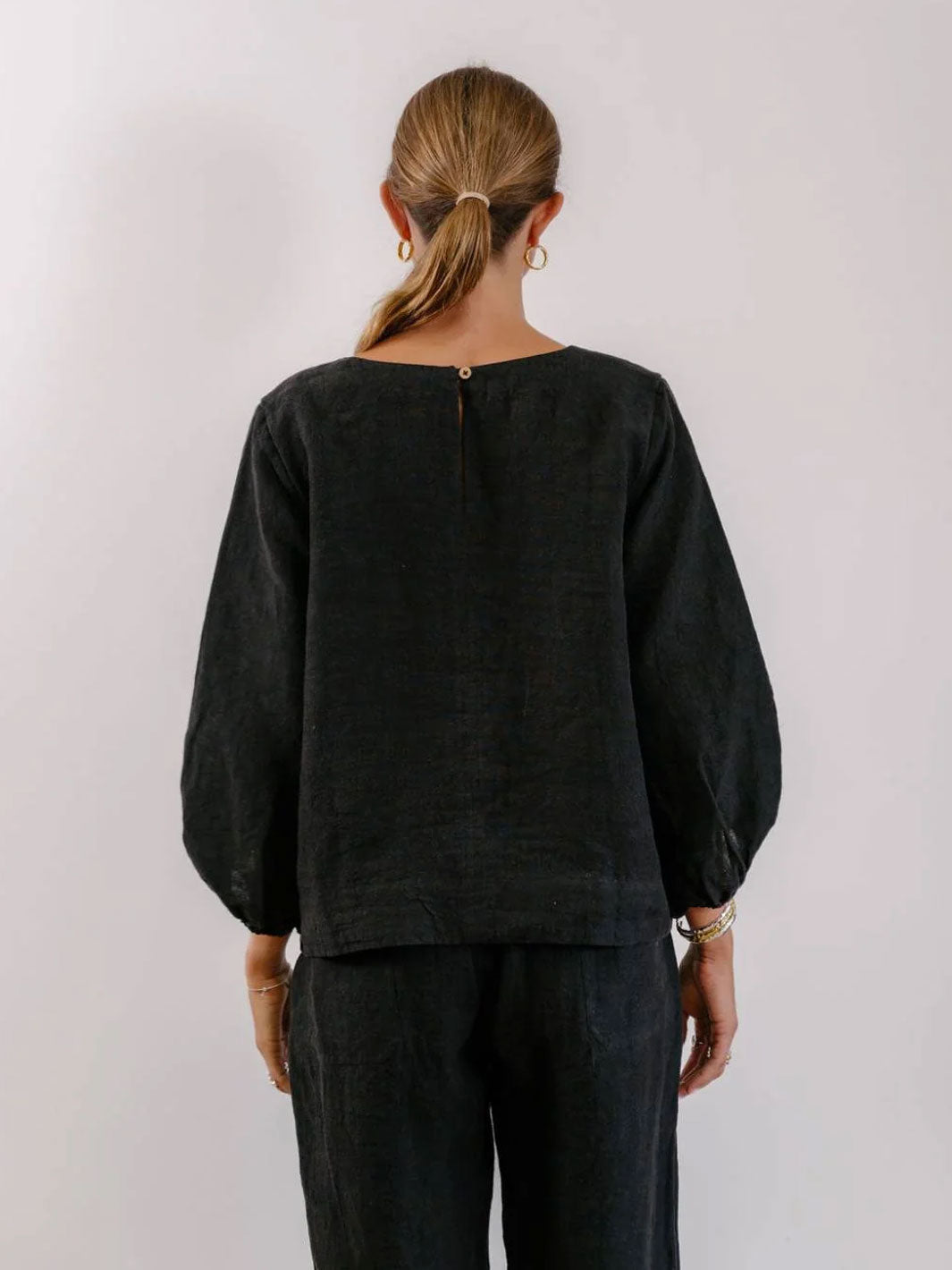 A woman viewed from the back wearing a loose black long-sleeve top with balloon sleeves and matching dark pants, standing against a plain light gray studio background.