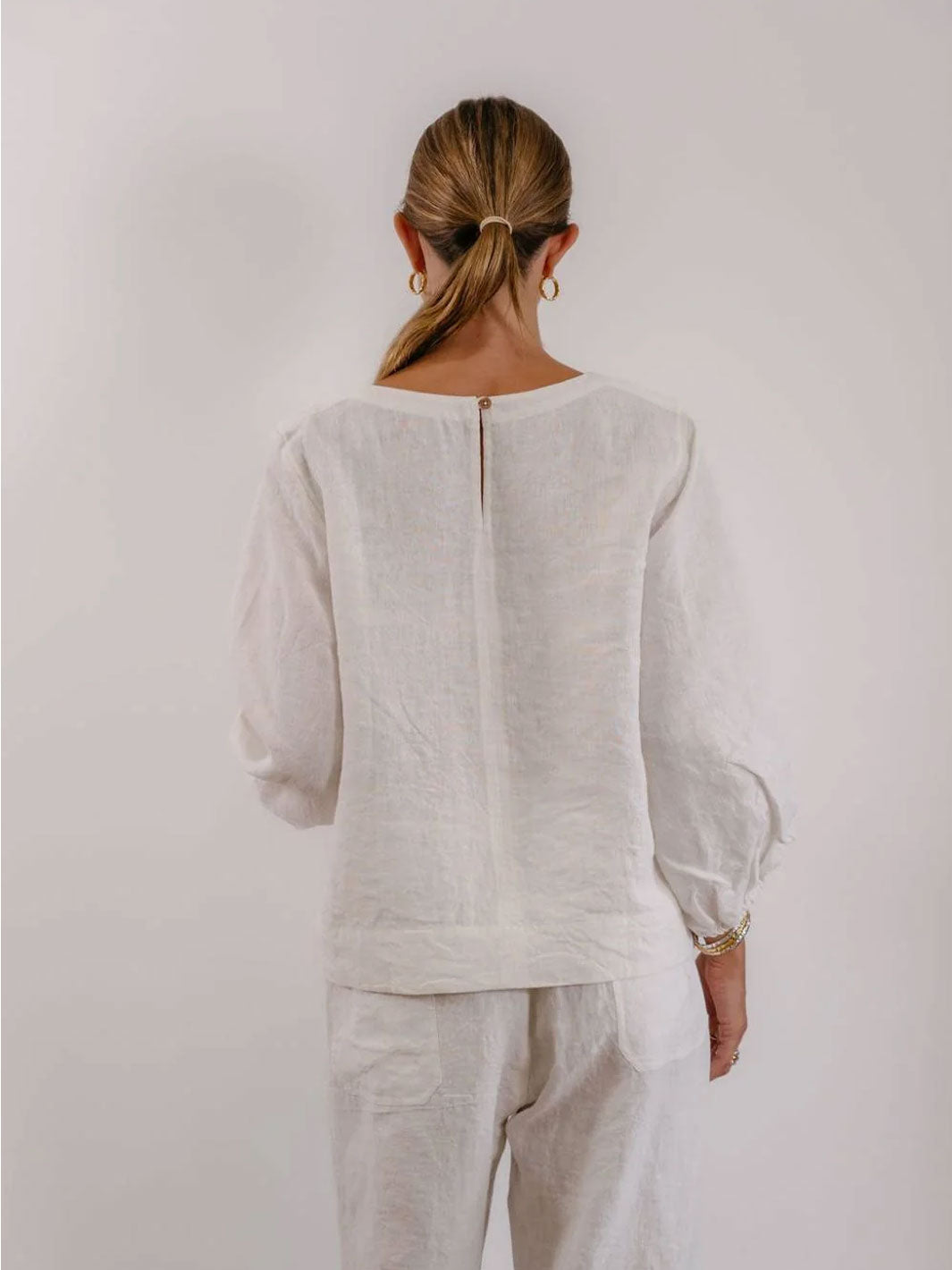A woman viewed from behind wearing a loose white linen top and matching white pants against a plain light background.