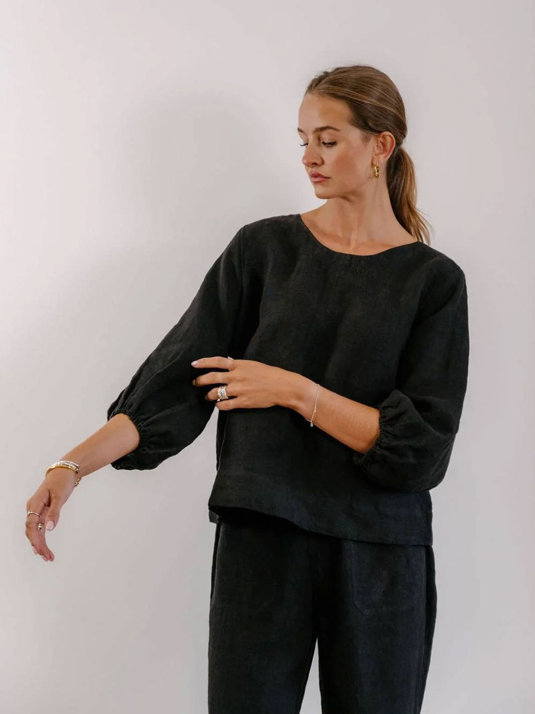 A woman wearing a black linen-blend top with voluminous balloon sleeves and matching dark trousers, standing against a plain light gray wall.
