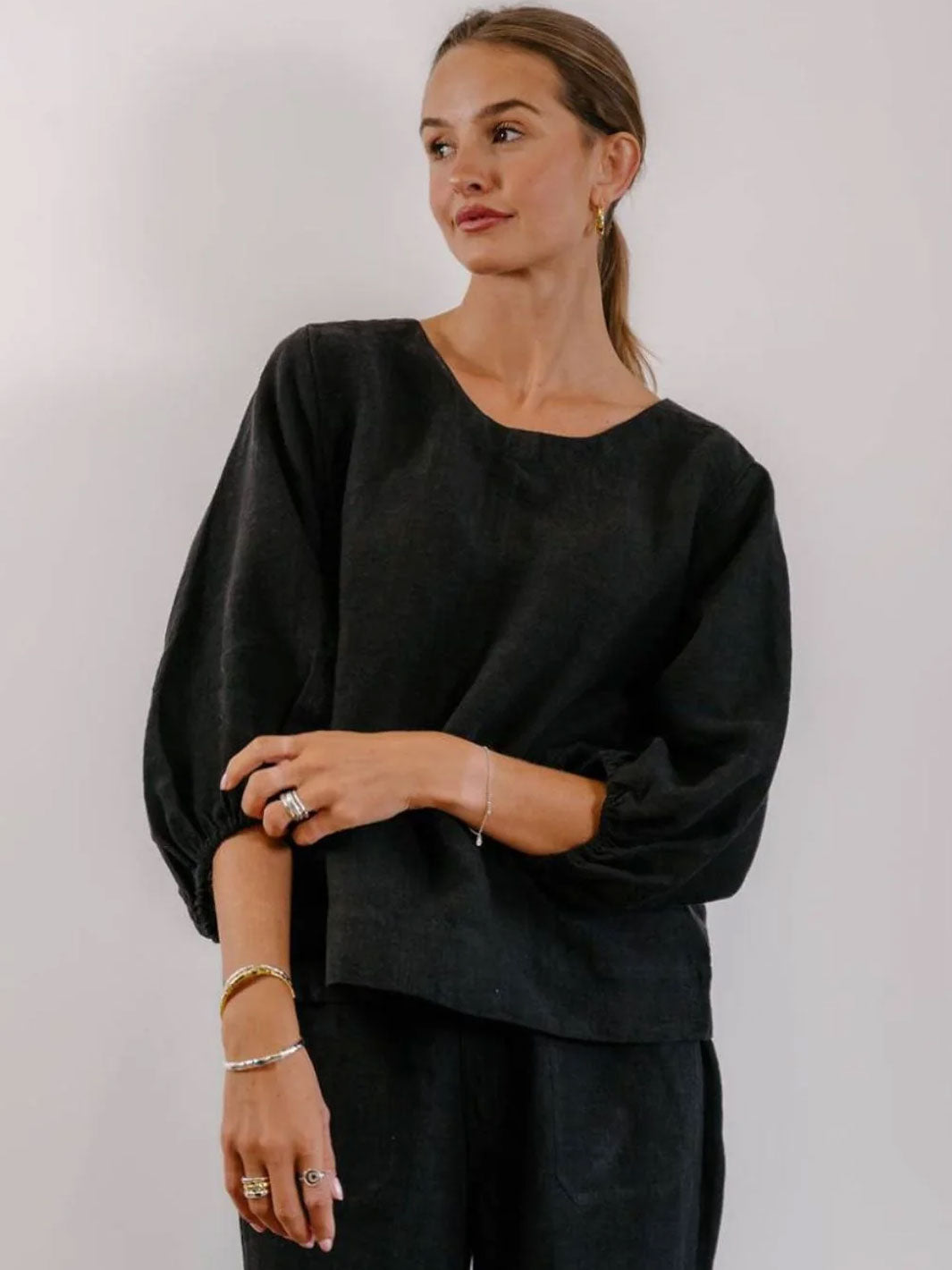 A woman wearing a loose black long-sleeve top with gathered elbow-length sleeves and black pants, standing against a plain light gray background.