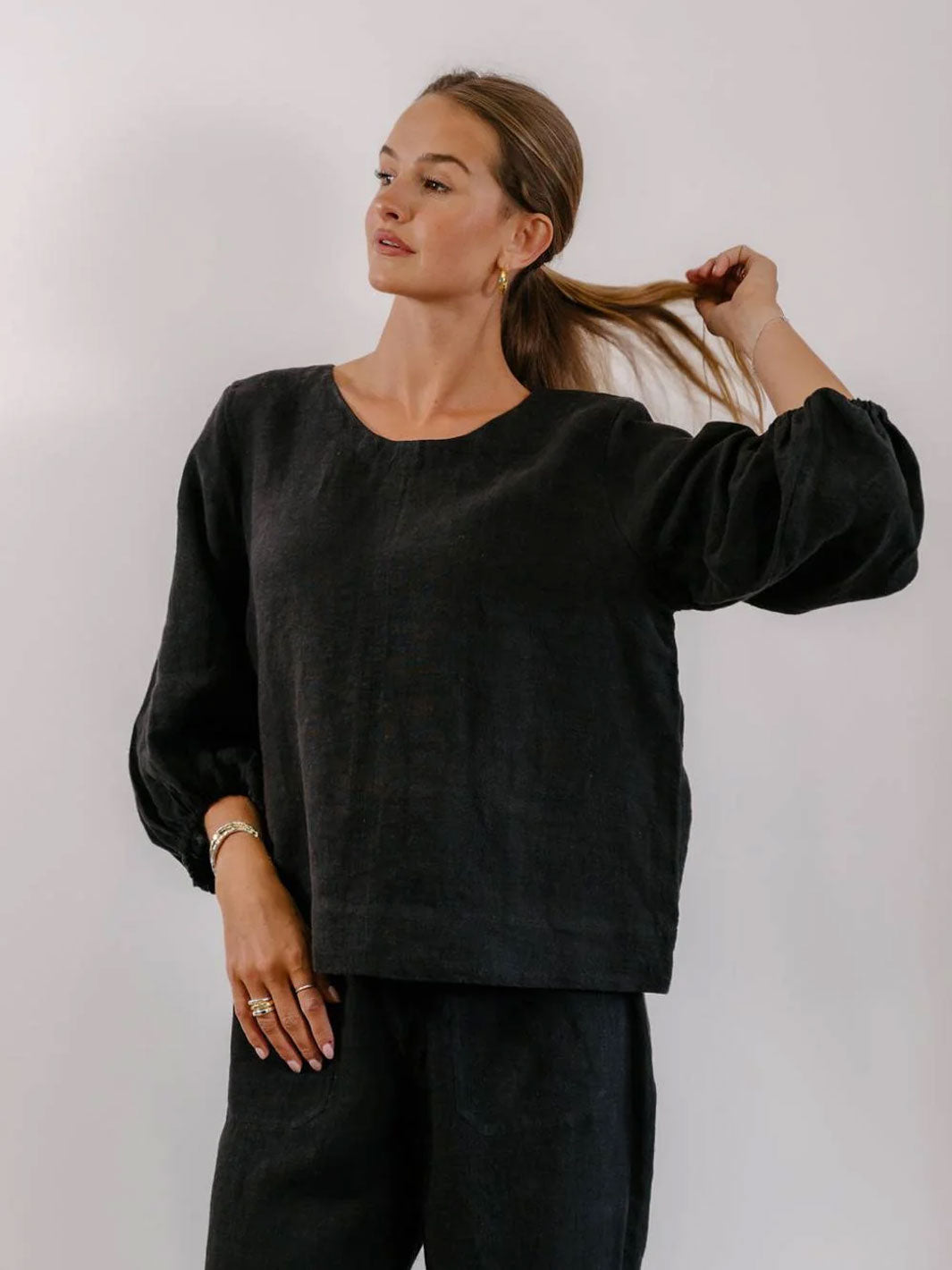 A woman wearing a loose black linen-blend top with voluminous long sleeves and matching dark pants, standing against a plain light gray studio background.