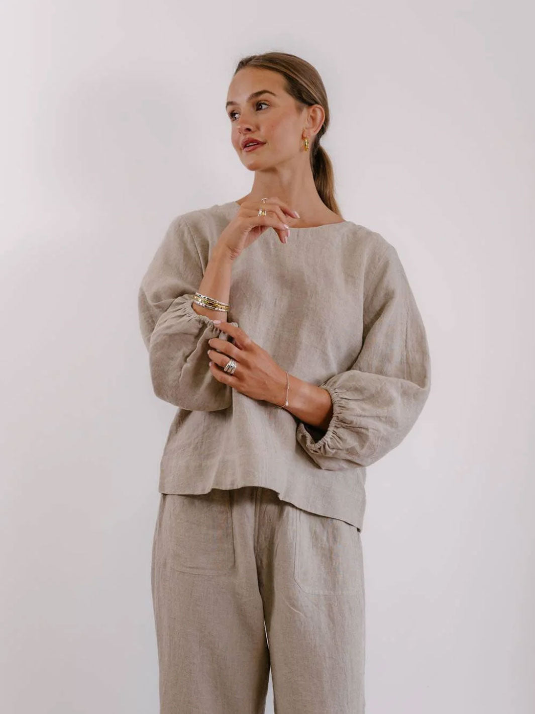 A woman modeling a light beige linen two-piece set consisting of a long-sleeve top with gathered cuffs and matching wide-leg trousers, set against a plain light gray background.