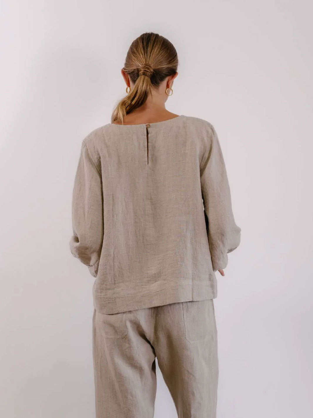 A woman viewed from behind wearing a light beige linen long-sleeve top and matching trousers against a plain light gray background.