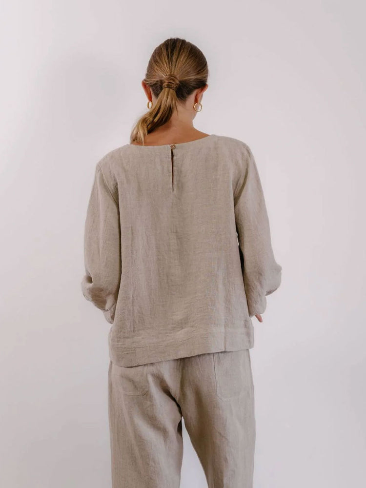A woman viewed from behind wearing a light beige linen long-sleeve top and matching trousers against a plain light gray background.
