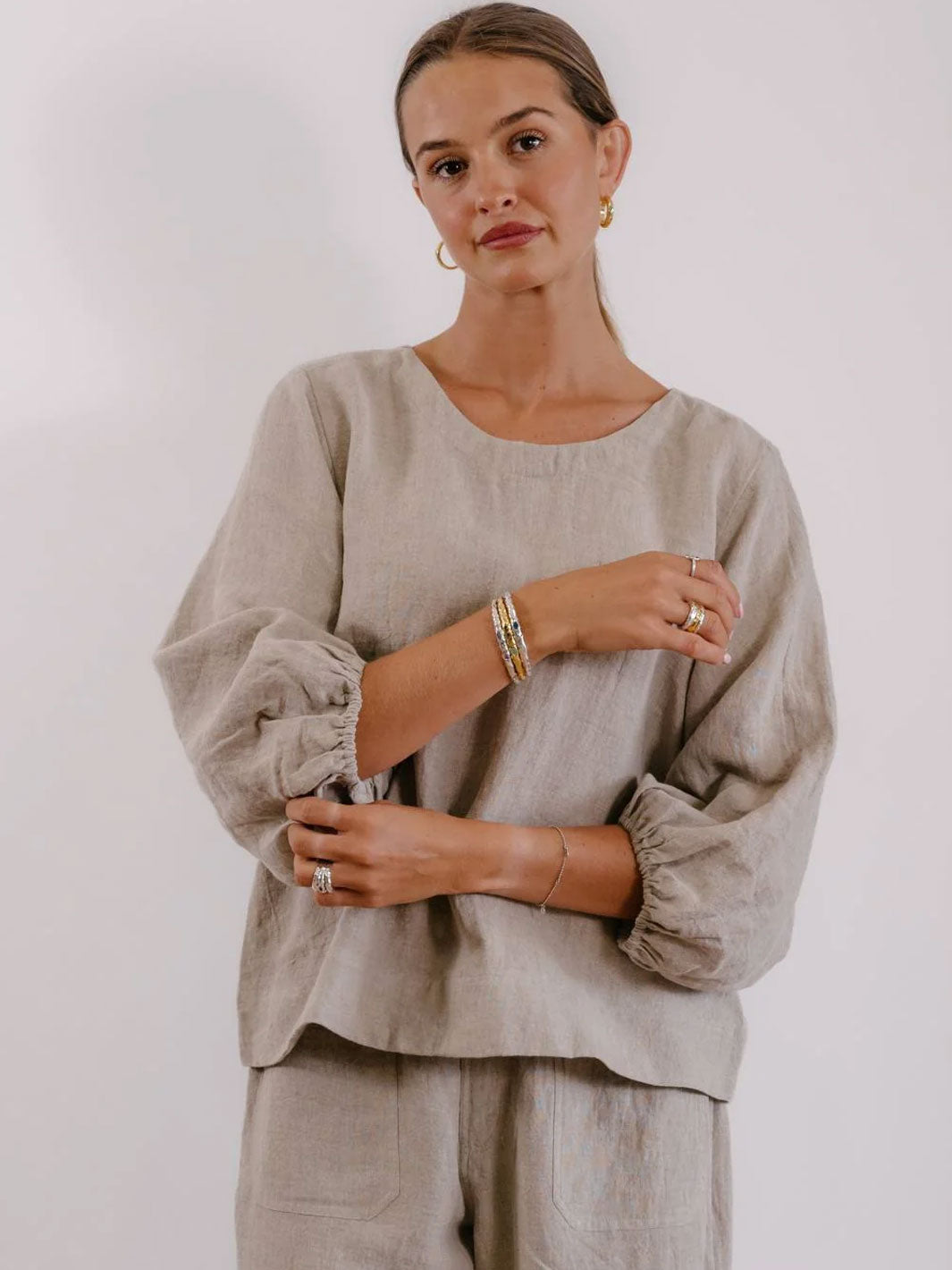 A woman wearing a light beige linen top with voluminous gathered sleeves and matching trousers, photographed against a plain light background.