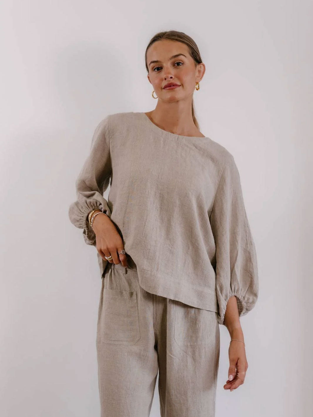 A woman modeling a light beige linen two-piece set consisting of a long-sleeve top with balloon sleeves and matching linen trousers, set against a plain light gray background.