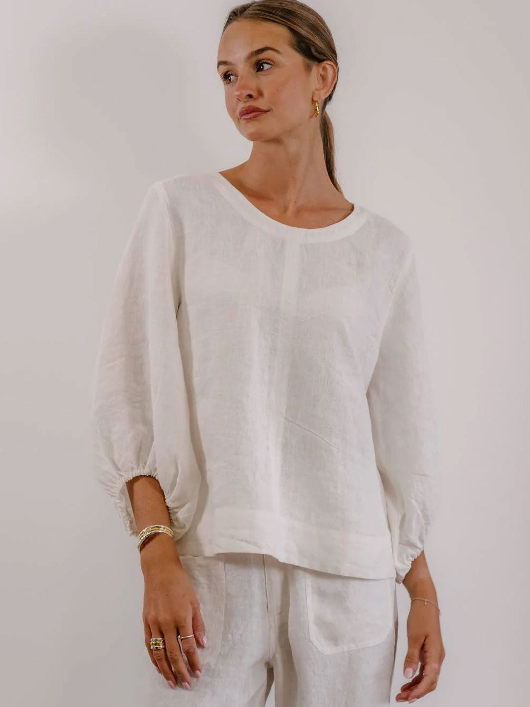 A woman wearing a loose white long-sleeve top with gathered sleeves and white pants, standing against a plain light background.