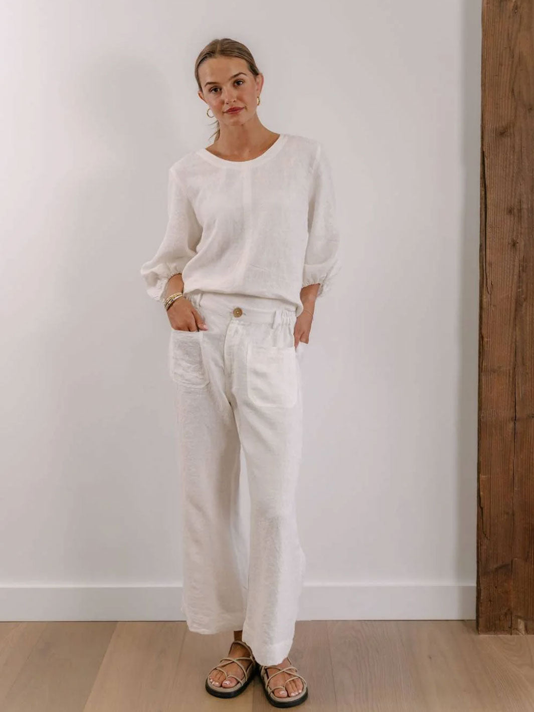 A woman modeling a white long-sleeve linen top and matching white wide-leg linen pants, paired with black sandals, standing against a white wall with a wooden column on the right.