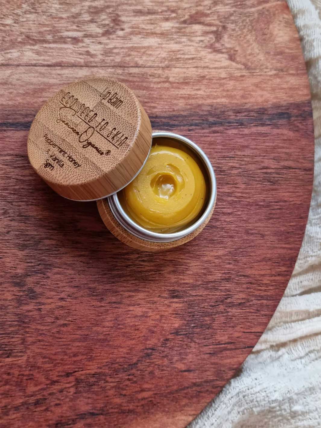 A small round metal tin with a bamboo lid, shown open with a yellow balm or wax inside, placed on a wooden surface. The lid is engraved with the brand name &