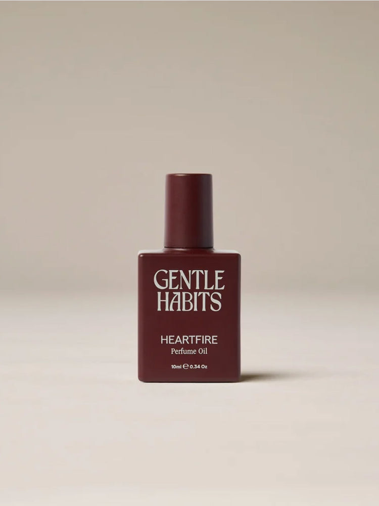 A bottle of Gentle Habits Heartfire Perfume Oil in a deep burgundy color, set against a neutral light beige background.