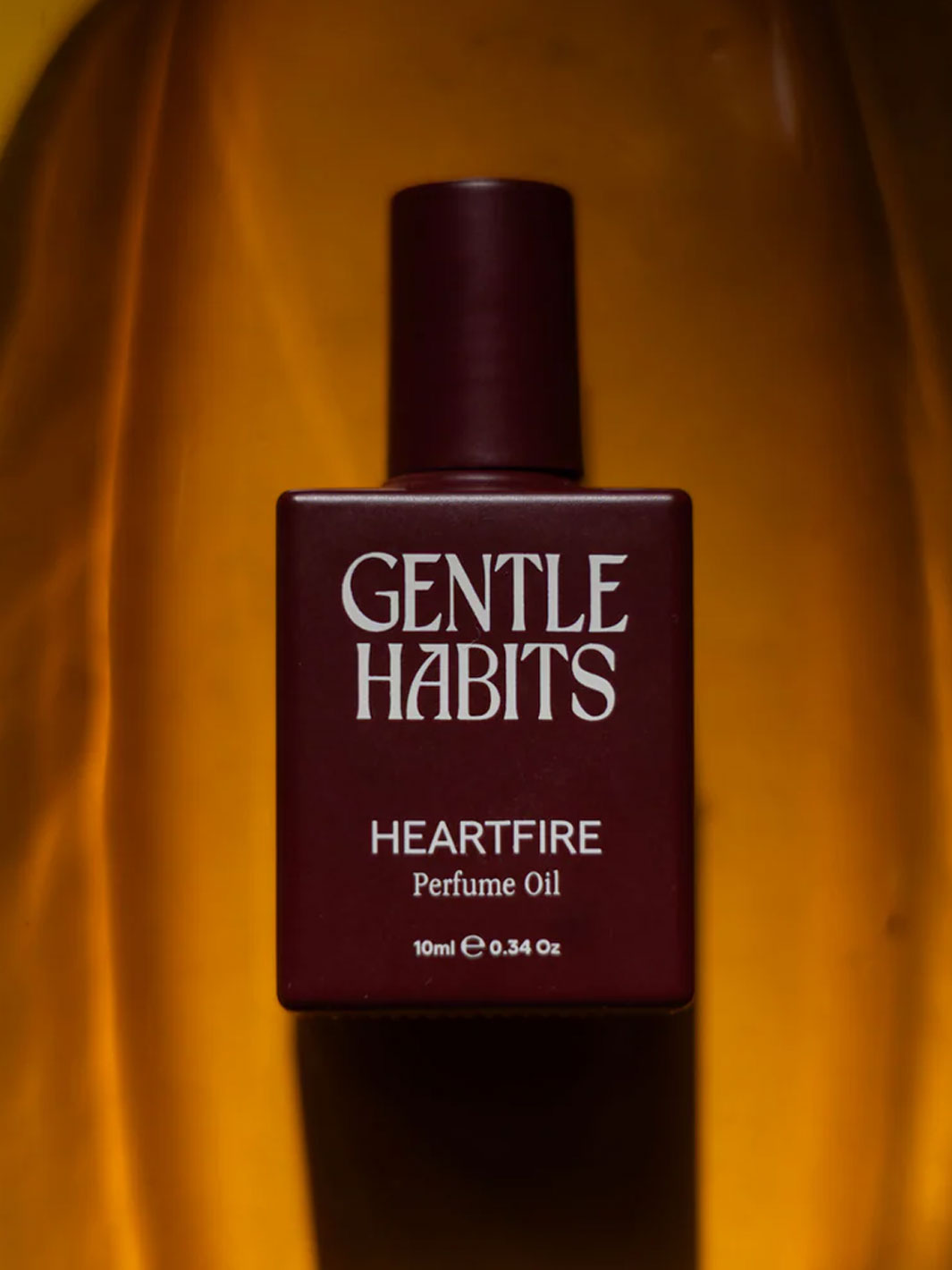 A bottle of Gentle Habits Heartfire Perfume Oil, featuring a dark burgundy rectangular bottle and cap with white text, set against a draped golden-yellow fabric background.