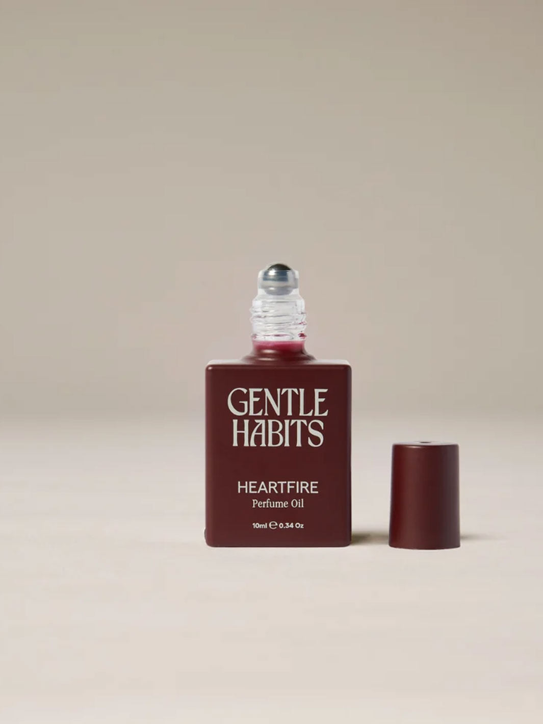A bottle of Gentle Habits Heartfire Perfume Oil with its cap removed and placed beside it, set against a solid neutral beige background.