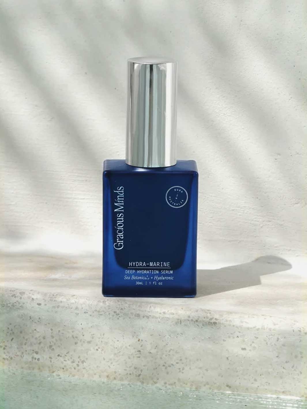 A bottle of Gracious Minds Hydra-Marine Deep Hydration Serum, featuring a deep blue rectangular bottle and a shiny silver cap, set against a textured light grey background with soft leaf shadows.
