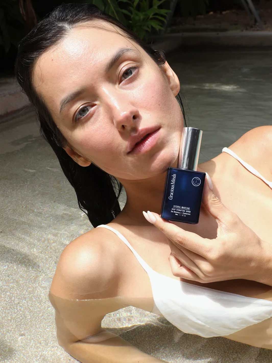 A woman in a white bikini holds a dark blue bottle of Gracious Minds facial serum against a poolside background.