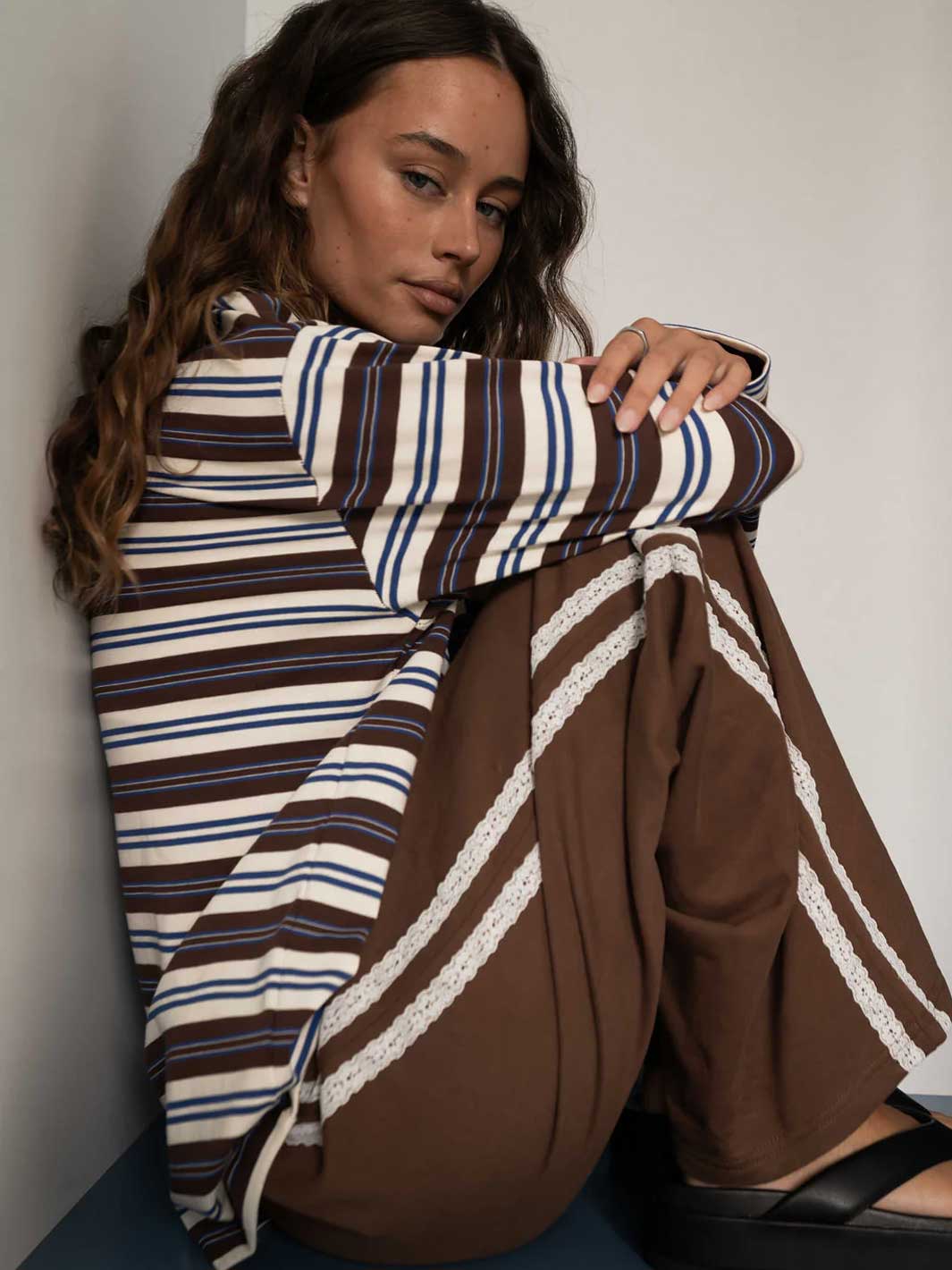 A woman sits against a plain wall wearing a brown, cream, and blue striped long-sleeve top and matching brown wide-leg pants with white lace trim.