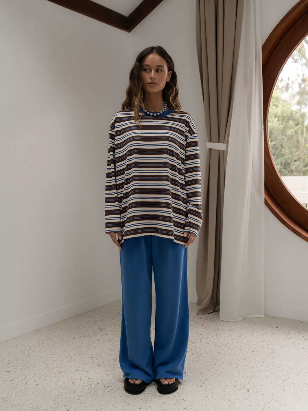 A woman stands in a minimalist room wearing a long-sleeve striped long sleeve t-shirt top with brown, blue, and cream horizontal stripes, paired with wide-leg blue pants and black sandals.