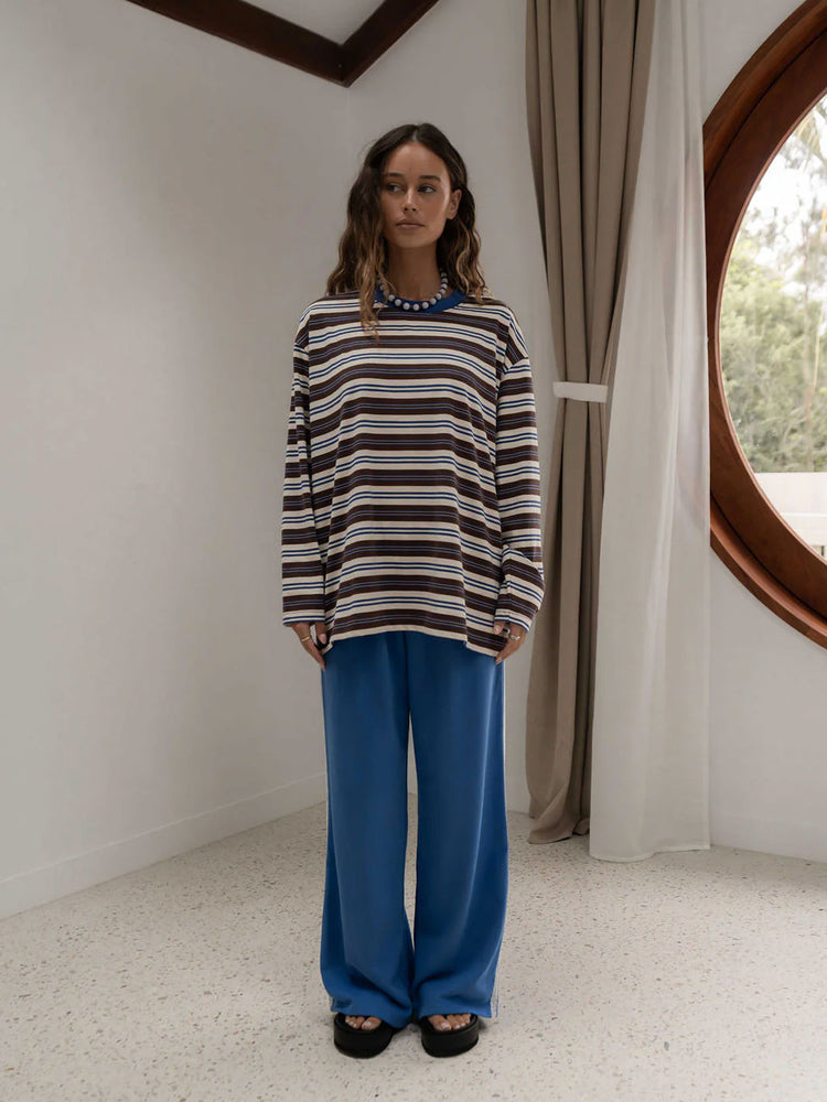 A woman stands in a minimalist room wearing a long-sleeve striped long sleeve t-shirt top with brown, blue, and cream horizontal stripes, paired with wide-leg blue pants and black sandals.