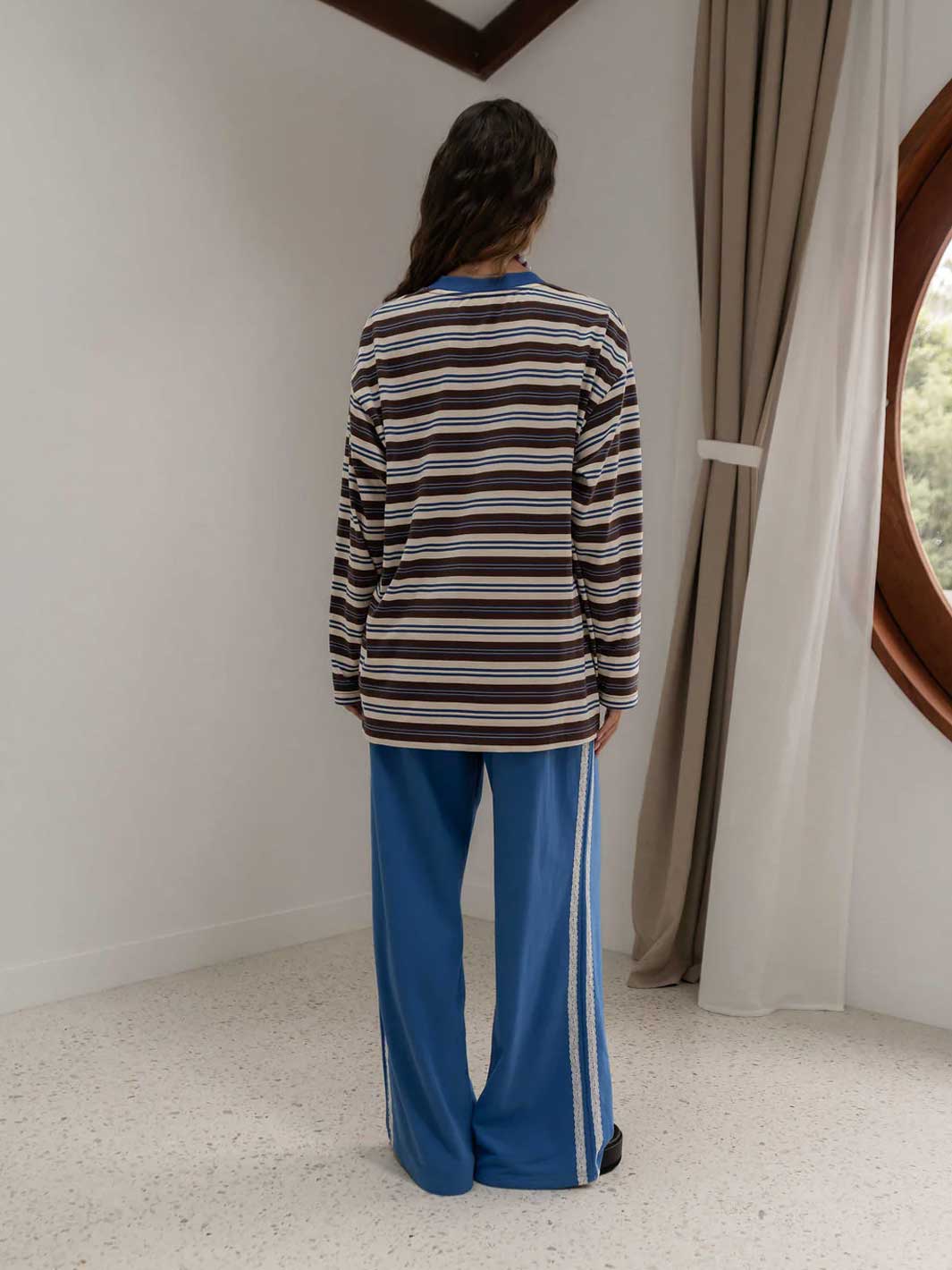 A person standing with their back to the camera, wearing a long-sleeve striped tee shirt with brown, blue, and cream horizontal stripes, paired with blue wide-leg pants featuring white side stripes.