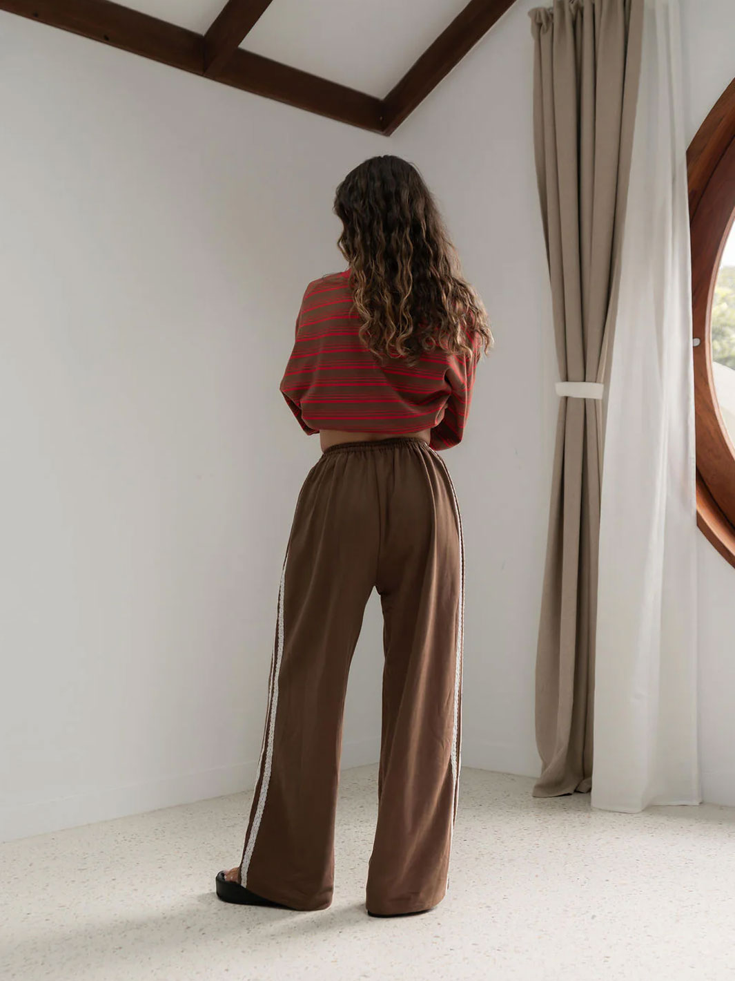 A woman stands with her back to the camera in a bright room with white walls, dark wood ceiling beams, and a window with beige curtains. She is wearing a rust-red and brown striped long-sleeve tee shirt top and brown wide-leg pants with white laces striped trim.