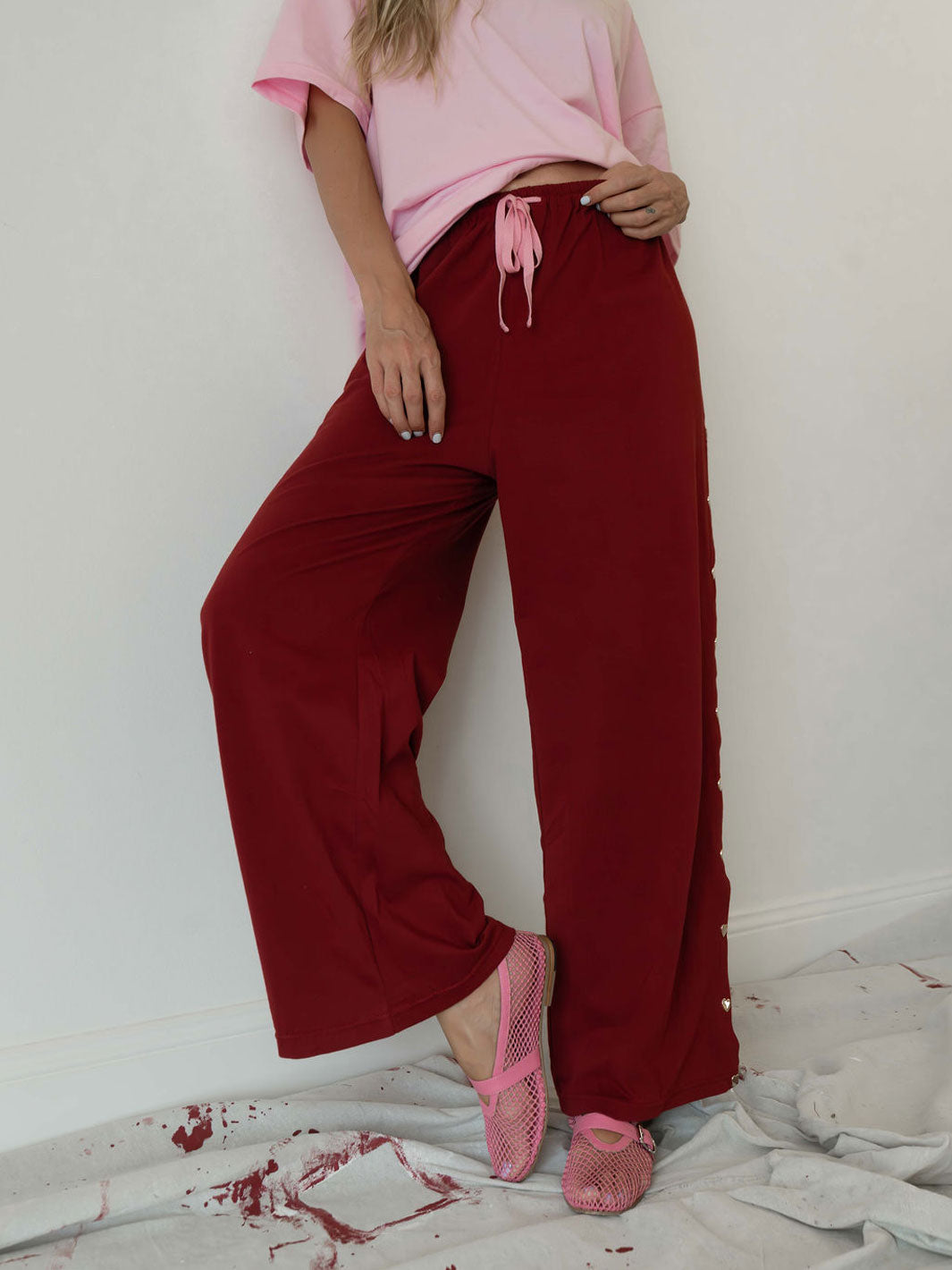 A model wearing loose, wide-leg burgundy pants with a pink drawstring waist, paired with a pink t-shirt and pink mesh flats, standing against a white wall.